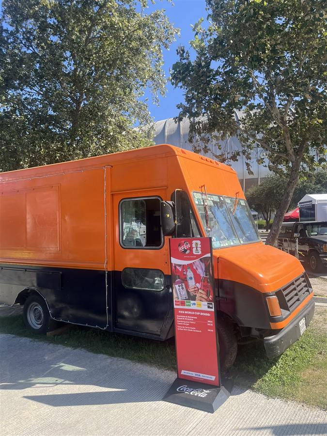Truck de Léon Foodtruck junto a un letrero oficial Coca-Cola FIFA World Cup 2026 con precios de Hamburguesa Clásica y Torta de Carne Asada